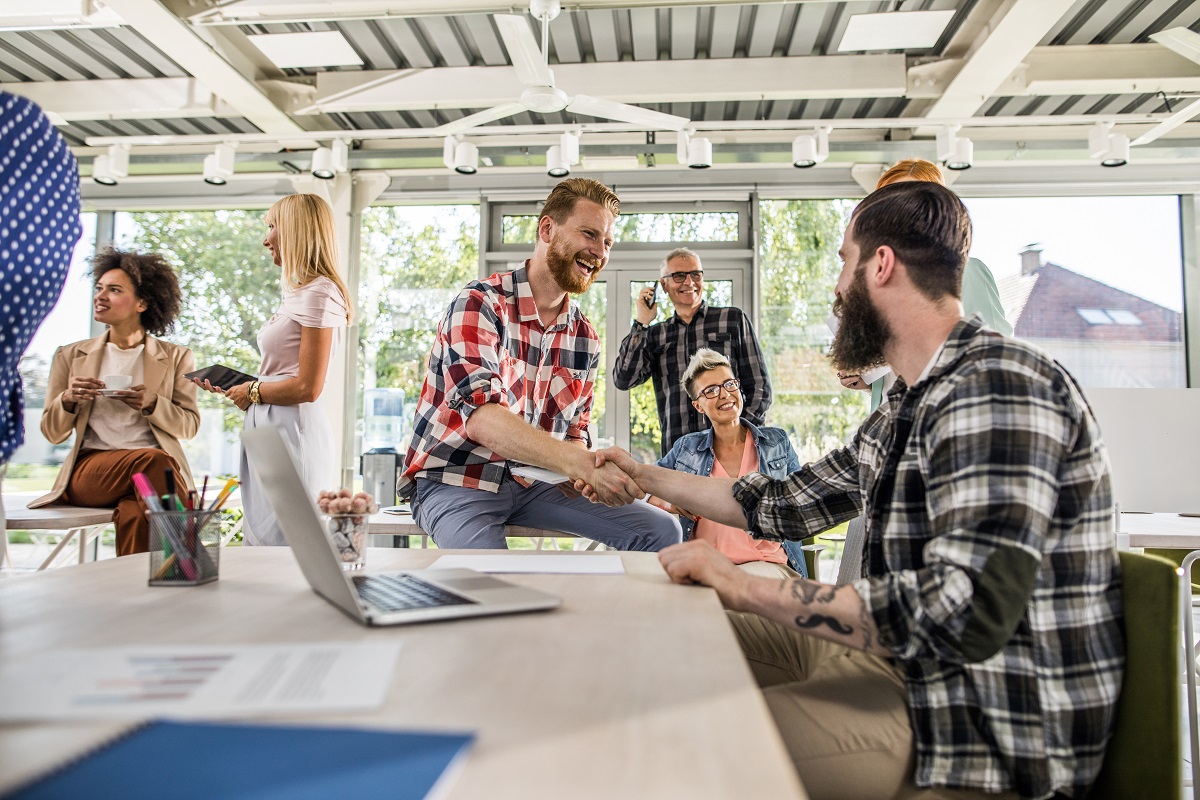 Cheerful businessman shaking hands with his colleague in a modern office full of people. Focus is on redhead man.